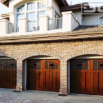 Three single-car garage doors in solid African Mahogany. Each custom garage door has an arched window top with glue chip glass panes divided with real mullions. At the center of each window there is a diamond shaped piece with clear glass. The bottom of each door is finished in long, vertical recessed panels. All three doors were stained in a natural walnut color to enrich the beauty of the wood.