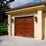 A single-car overhead garage door in a French Provincial style. The wooden garage door boasts an exquisite European style with a faux arch top detail. The top third of the door consists of two hand-forged scrolls that were beautifully inlaid into the wood. The mid third section is composed of vertical tongue and groove planks that simulate French bead board and the bottom third of the garage door is accentuated with diamond trim work that is reminiscent of French architecture. A set of custom levers give the door the total effect of out-swing carriage doors!