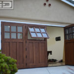 Seen in this image is a double car garage door conversion. The door configuration consists of a four panel bi-folding configuration that parts in the middle with two panels folding out to the right and two panels bi-folding to the left. Each bi-folding panel boasts a functional awning window at the top with a six glass pane configuration, truly divided by mullions. The open window shows a bug screen that will keep bugs out when open but let ventilation into the converted garage. The bottom panel of the system is composed of three vertical recessed panels that give this bi-folding carriage door configuration its Craftsman Architectural Style. To accentuate the beauty of the clear alder wood grain we stained the door in a warm, golden color similar to natural cedar wood.