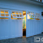 Custom architectural out-swing carriage doors in white with functional, awning windows. Handcrafted in composite materials with clear glass windows with six panes divided by true mullions. Two large door panels swing out on this double car garage door opening.