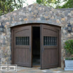 Pictured in this image is a Tuscan style carriage garage door in a two-leaf out-swing format. The carriage doors are slightly arched at the top to match the garage opening. The carriage door panels are divided into three sections with the top quarter consisting of vertical tongue and groove planks in variant widths. Below that section you'll see a quarter of the panel with a large window fitted with antique seedy glass and vertical iron balusters. The bottom half of the door panels is, again, composed of vertical tongue and groove planks in variant widths to match the very top of the panels. The carriage doors are handsomely crafted out of rustic alder wood with an oil rub finish which gives the automatic carriage doors an old world look with the convenience of modern day automation. These doors can be opened at the push of a button!