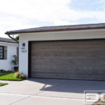 In this photograph, there is a modern farmhouse residence with a reclaimed wood double-car garage door and matching shutters. The double-car garage door boasts a minimalist design of horizontal-oriented reclaimed wood planks. The simple design of the door allows the eye to admire the natural character of the aged wood’s surface with all its naturally carved grain structure and color tonalities. The rustic surface of the minimalist garage door is a wonderful contrast against the crisp white walls of this modern farmhouse. To complement the garage door and complete the entire curb appeal of this home, several decorative window shutters created out of the same lumber used on the garage decorate many of the windows on this home.